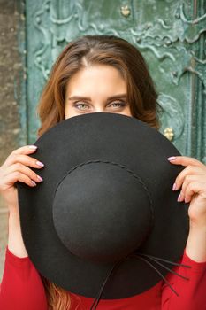 A fashionable beautiful young caucasian woman hiding face with a round black hat on the background the ancient door