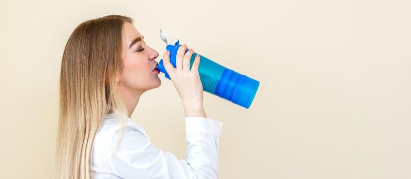 Side view of beautiful young caucasian woman is drinking water from plastic bottle against a beige background