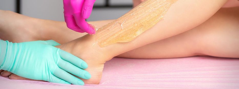 Two female cosmetologists waxing woman's legs with hot sugar paste in a beauty spa salon