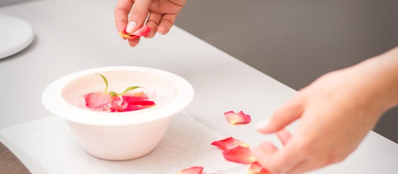 Beautician's female hands preparing manicure bath with red and pink roses petals on the table in spa