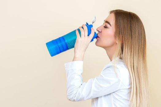 Side view of beautiful young caucasian woman is drinking water from plastic bottle against a beige background