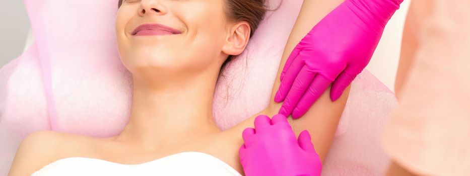 Beautiful young caucasian smiling woman receiving waxing her armpit by hands of a beautician in a medical spa