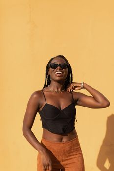 Portrait of a young black female posing with style and smiling with a plain orange wall behind her during a summer sunset