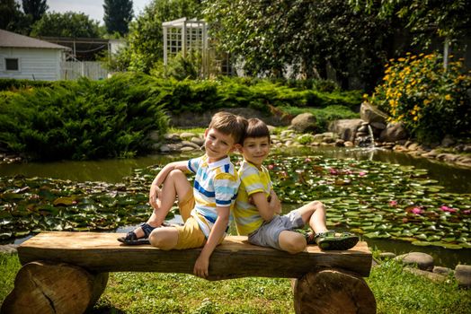 Two brother boys sitting on a bench in a park near peaceful lake with water lily Nymphaeum. Kids relaxing on nature on hot summer day.