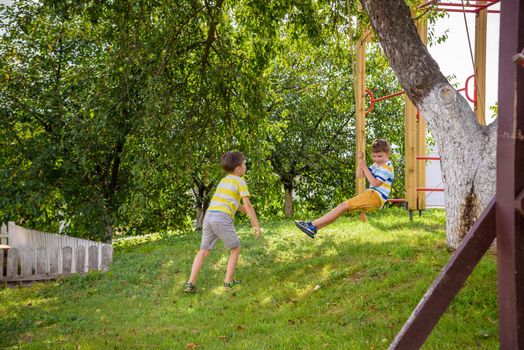 Two adorable happy little boys is having fun on a rope swing which he has found while having rest outside city. Active leisure time with children.