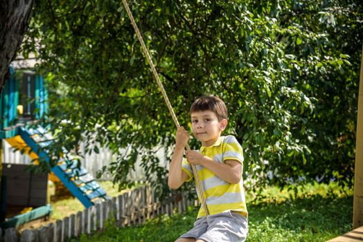 Happy little boy is having fun on a rope swing which he has found while having rest outside city. Active leisure time with children.