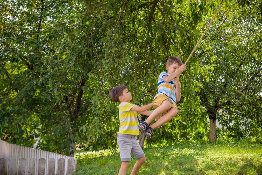 Two adorable happy little boys is having fun on a rope swing which he has found while having rest outside city. Active leisure time with children.