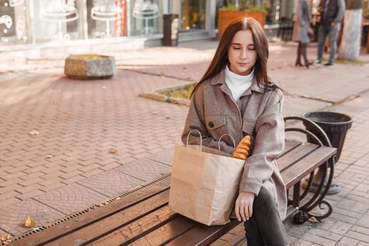 A stylish brunette woman walks around the autumn city. The brunette is sitting on a bench outside with a glass of coffee and a phone, holding a craft bag with a baguette.