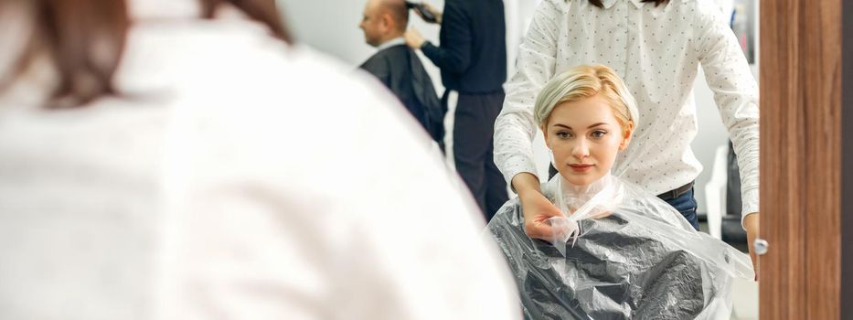 The female hairdresser puts on a transparent cellophane cape to the female client in a beauty hair salon