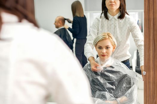 The female hairdresser puts on a transparent cellophane cape to the female client in a beauty hair salon