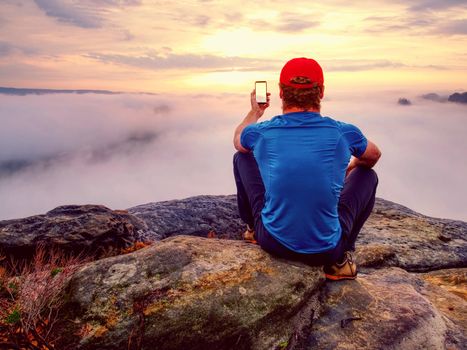 Courageous man sitting on the peak of rock makes the photo on the his phone. Autumn nature 