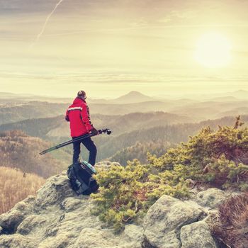 Nature photographer with tripod and camera on cliff and thinking. Dreamy fogy landscape orange misty sunrise in a beautiful mountains
