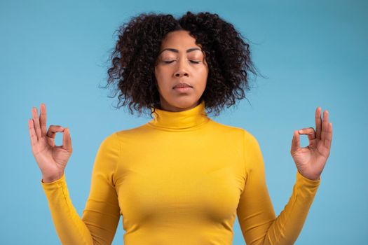 Calm african woman in blue relaxing, meditating. She calms down, breathes deeply with mudra om on blue studio background. Yoga concept. High quality photo