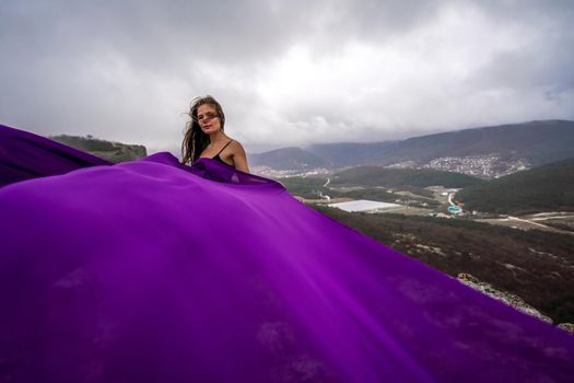 A woman with long hair is standing in a purple flowing dress with a flowing fabric. On the mountain against the background of the sky with clouds