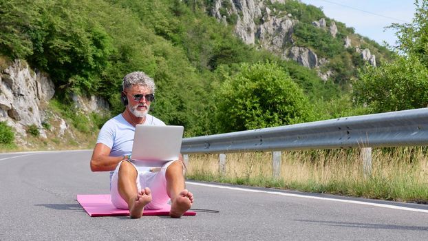 Gray-haired elderly man freelancer with a beard in sunglasses, working on a laptop on nature, road, mountains. A crazy and extraordinary old man sits on a yoga mat in the summer