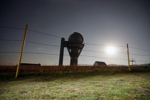 A big bright moon in the starry sky. The observatory is fenced with a barbed fence. The dome above the building with a large telesk. The yellow-green grass is trampled. There are several houses