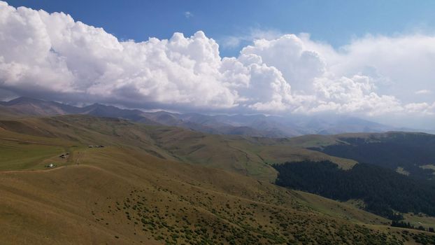 Big white clouds over green hills and mountains. Top view from the drone on endless fields. Roads are visible in places, herds of animals graze. A tent camp has been set up. Coniferous trees in gorge