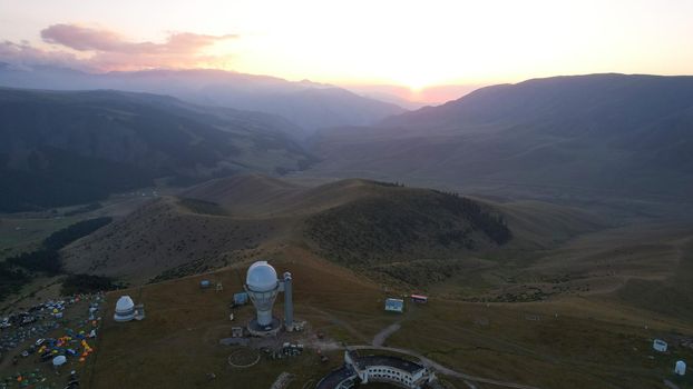 Two large telescope domes at sunset. Drone view of Assy-Turgen Observatory. Beautiful red sunset. Green hills and clouds. Tourists watch the sun. There is a large tent camp and cars nearby. Kazakhstan