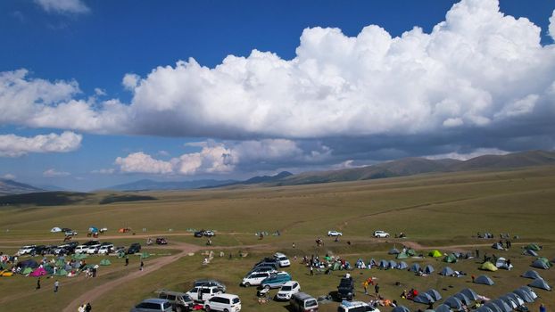 Big white clouds over green hills and mountains. Top view from the drone on endless fields. Roads are visible in places, herds of animals graze. A tent camp has been set up. Coniferous trees in gorge