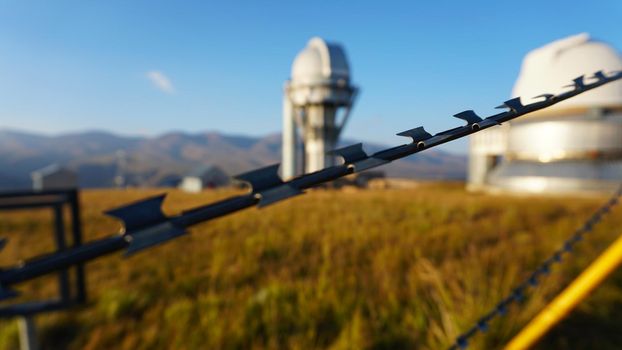 A large observatory is protected by a barbed fence. There is a long sharp wire around perimeter. Buildings with domes and a telescope are visible in distance. Yellow-green grass. Mountainous terrain