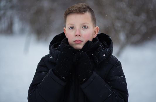 A cute boy of 13-15 years old in dark winter clothes and in black gloves in an open space looks at the camera.