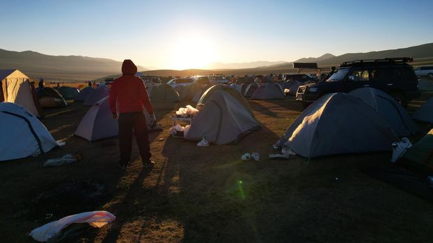 A guy at dawn looks at a camp covered in garbage. The bright sun illuminates tents, hills and fields. The shadow of tents fall on ground where the garbage lies. Bags, shoes, and paper are scattered