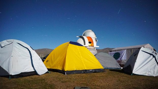 A camp with colored tents near the observatory. The Perseid starfall. A large teleskom watches the starry night sky. Meteorites and planes fly by, leaving traces in the sky. The stars are burning.