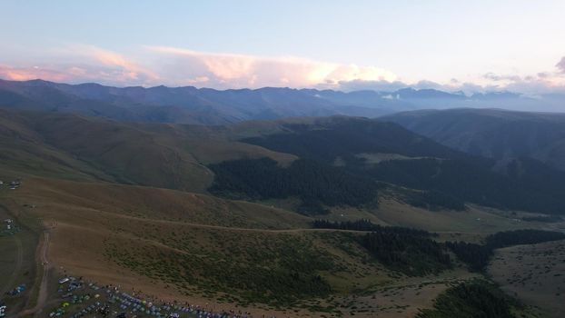 Red rays of the sun at sunset among the mountains. Green grass turns yellow, in places a small forest. Tourists walk in the hills. A river runs in the distance. The clouds are highlighted in pink.