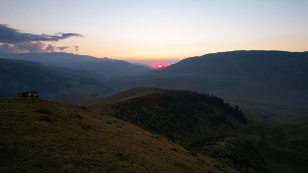 Red rays of the sun at sunset among the mountains. Green grass turns yellow, in places a small forest. Tourists walk in the hills. A river runs in the distance. The clouds are highlighted in pink.