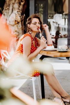 Charming woman in a restaurant, cafe on the street. She sits at the table and eats a cake with a fork. Dressed in a red sundress with white polka dots