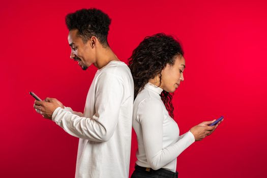 Young african couple stand with their backs to each other and print messages or surf the Internet from their mobile phone. Technology, smartphone, social networks concept.