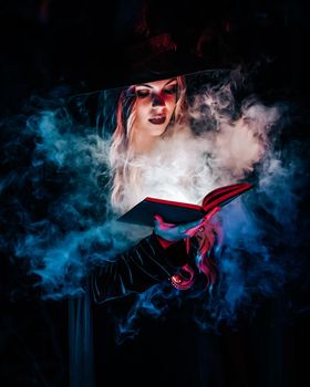 Beautiful witch reading magic book on dark background in forest. Halloween, blonde wizard costume concept. Pretty woman in blue smoke. . High quality photo