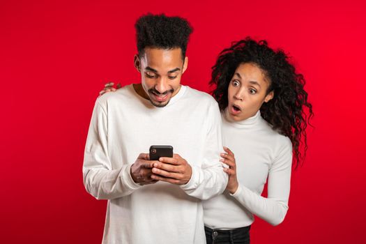 Pretty african girl is peeking over her boyfriends shoulder, he is communicating via mobile telephone. Woman shocked by jealousy and betrayal of her husband. Red studio background