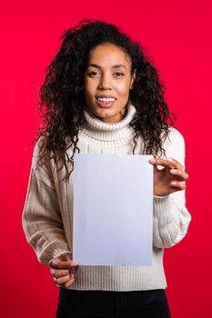Pretty mixed race girl with afro hair holding white a4 paper poster. Copy space. Smiling trendy woman on red studio background