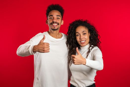 Young african american couple making thumbs up like sign over red background. Winner. Success. Positive girl and man smiles to camera. Body language