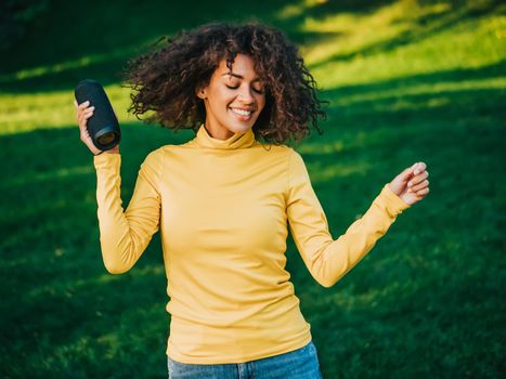 Modern trendy african american girl listening to music by wireless portable speaker.Young beautiful woman enjoying, dancing in park