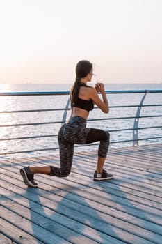 Young athletic girl performs squats on wooden embankment by the sea in early morning. Healthy lifestyle, coaching, training concept
