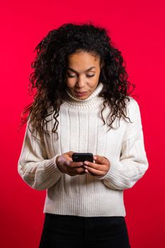 Girl with afro hair using smartphone, surfing internet or playing game on red studio background. Modern technology - apps, social networks.