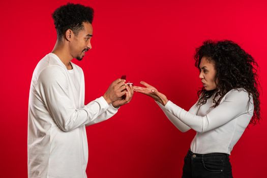 Young couple. African man makes marriage proposal to his lover woman with ring on red studio background but disappointed upset girl doesn't like gift