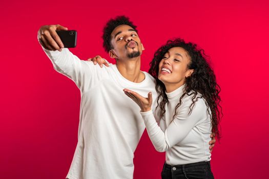 Young african american couple in white making selfie with smartphone on red studio background.Lovers match, love, holidays, happiness concept.