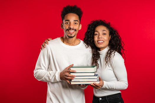 African students on red background in studio holds stack of university books from library. Classmates smiles, they happy to graduate