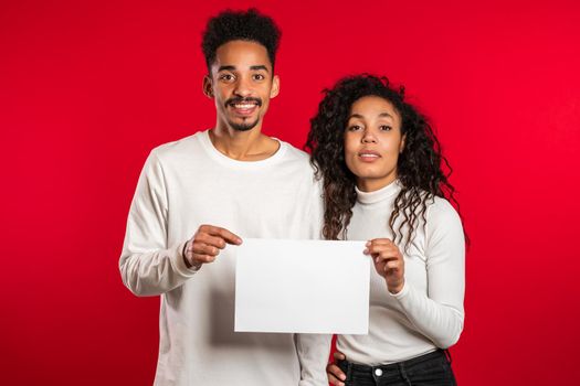 Portrait of young handsome african american man and woman couple holding white empty paper blank on red studio background. Copy space. Match with sign.