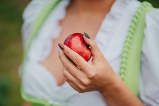 Woman in dirndl offers red apple as symbol of temptation, poison. Lady holding in hand ripe fruit. Fairy tale concept, halloween, cosplay, harvest. High quality photo