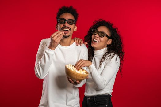Young couple in cinema in 3d glasses watching fascinating comedy movie and eating popcorn on red studio background