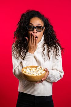 Young african woman in 3d glasses watching horror movie and eating popcorn on red studio background. Frightened girl with curly hair because of unexpectedness plot in film