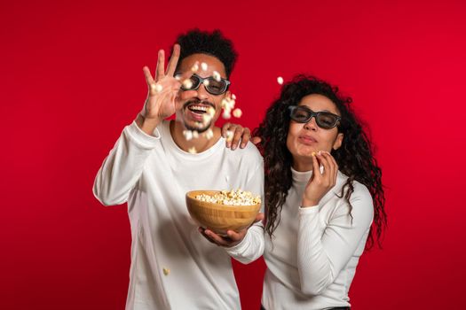 Young couple in cinema in 3d glasses watching fascinating comedy movie and eating popcorn on red studio background