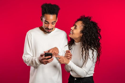 Young couple using their mobile devices, laughing and talking about it. Social apps on red studio background.