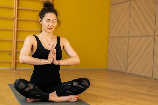 Young woman in black practicing yoga at light minimalist studio class room. Girl sitting in lotus pose and doing namaste. Health lifestyle concept. High quality photo