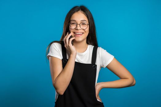 Pretty asian girl with long hair speaks on phone and laughs infectiously from interlocutor's jokes. Trendy outfit. Blue studio background.