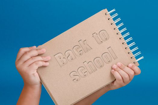 Notepad in hand with text back to school. Notepad in the hands of a child on a blue background. Hands hold a book as a symbol of the beginning of the school year and the day of knowledge